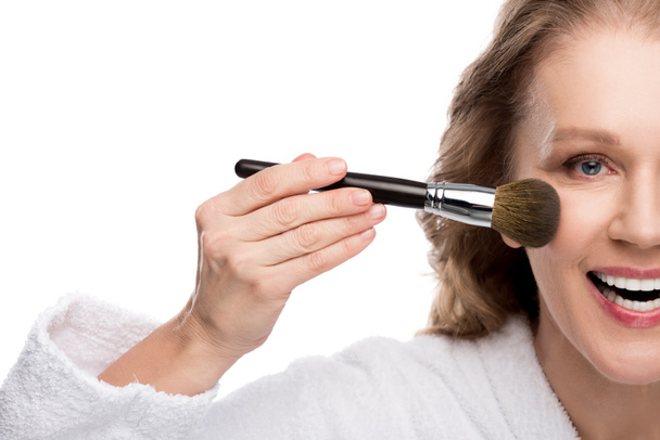 bijgesneden weergave van volwassen vrouw in badjas met cosmetica borstel tijdens het aanbrengen van make-up geïsoleerd op wit - Foto, afbeelding
