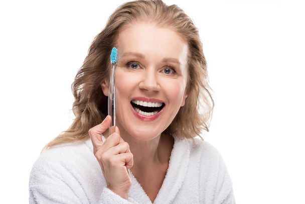 χαμογελαστή μεσήλικη γυναίκα σε μπουρνούζι με οδοντόβουρτσα απομονωμένη σε λευκό  - Φωτογραφία, εικόνα