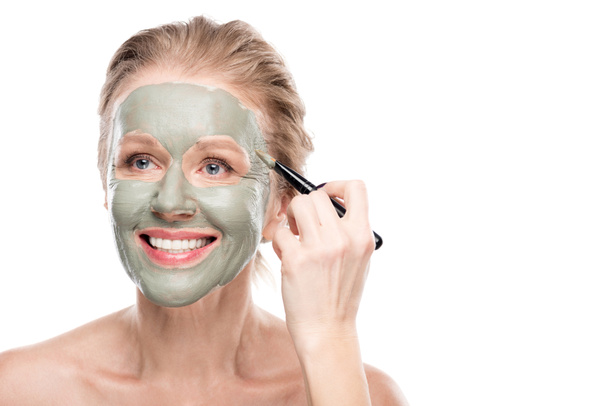 mujer madura sonriente Aplicando máscara de arcilla con cepillo cosmético aislado en blanco
 - Foto, Imagen