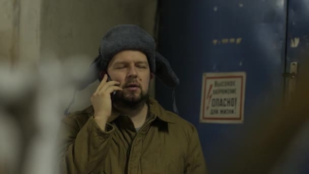 Russische hacker in de hoed met oorkleppen spreekt aan de telefoon in de kelder. Een man van dichte build. Russische genie. - Video