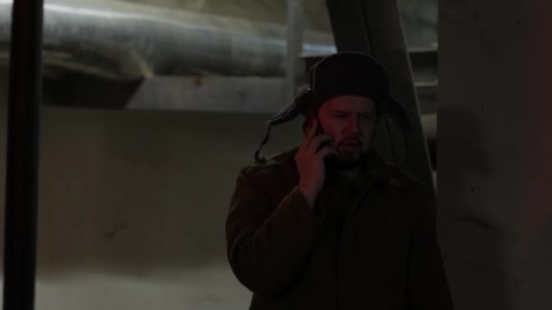 Liga a lâmpada do tecto. Hacker russo no chapéu com abas de ouvido fala ao telefone na cave. Um homem de construção densa. Genial russo
. - Filmagem, Vídeo