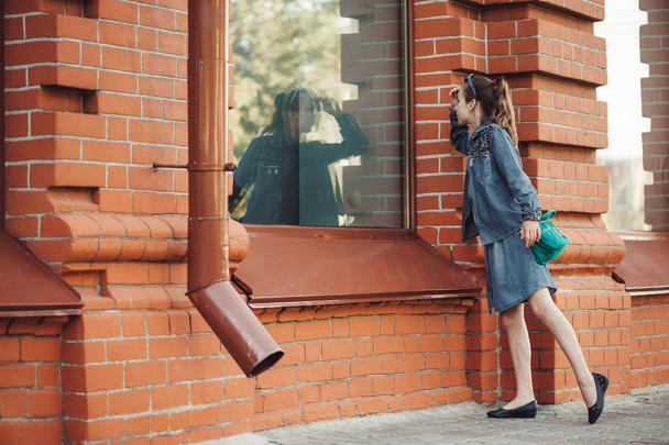Teenage girl in blue denim dress look in window of building from red (brown) brick outdoors. Young serious child piddle near shop. Reflex of dreamer in big window. Girl wants to do shopping. - Photo, Image