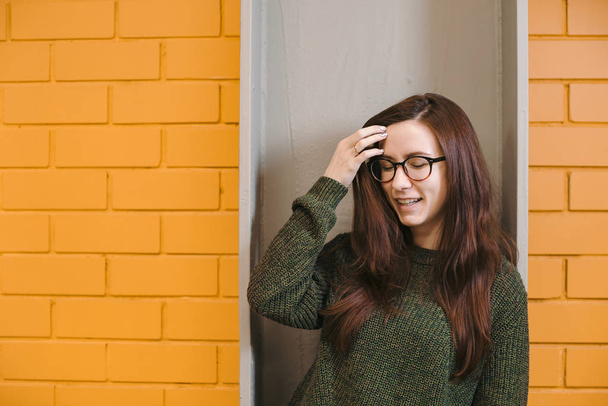 Красивая рыжая девушка в очках и зеленый свитер улыбается на фоне желтой кирпичной стены
. - Фото, изображение