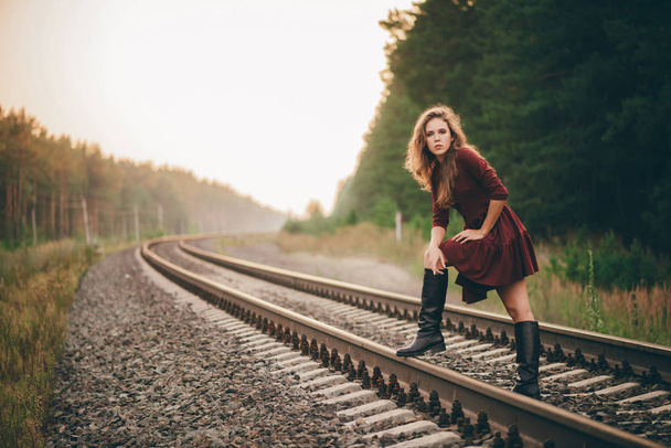 Beautiful dreamy girl with curly natural hair enjoy nature in forest on railway. Dreamer lady in burgundy dress walk on railroad. Inspired girl on rails at dawn. Sun in hair in autumn. Good mood. - Фото, изображение