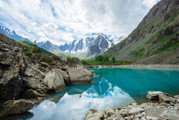 Mountain lake is surrounded by large stones and boulders on front of giant beautiful glacier. Amazing snowy mountains. Ridge with snow. Wonderful atmospheric landscape of majestic nature of highlands. - Photo, Image