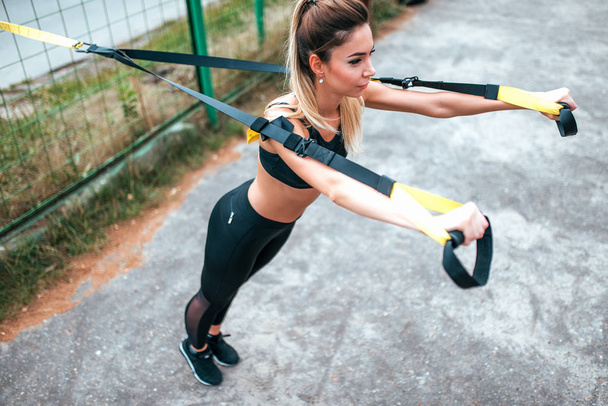 Girl in sportswear leggings and top. It is wrung out on loops for training by fitness. In summer in city near fence. Tanned skin is beautiful figure. Push-ups on straps. Active lifestyle in the city. - Foto, Imagem