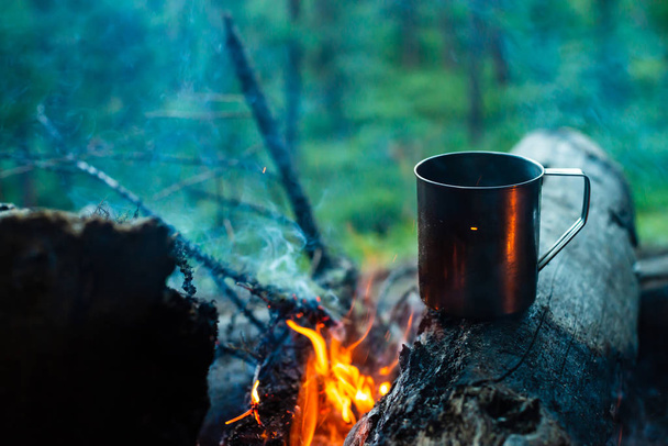 Tea in metal mug heats up on stone on bonfire. Hot drink on nature. Tea drinking in open air. Camping in dusk. Romantic warm atmosphere outdoor in twilight . Active rest. - Photo, Image