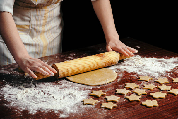 Uma mulher na cozinha está rolando uma massa em uma mesa de madeira com um rolo de madeira. Ela molda biscoitos na forma de asteriscos
. - Foto, Imagem