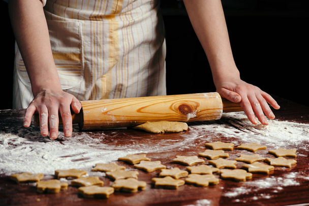 Женщина на кухне катит тесто на деревянный стол деревянной скалкой. Она делает печенье в форме звездочек.
. - Фото, изображение