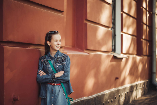Cheerful little girl in jeans clothes stands near an old restored beautiful building. - Фото, изображение