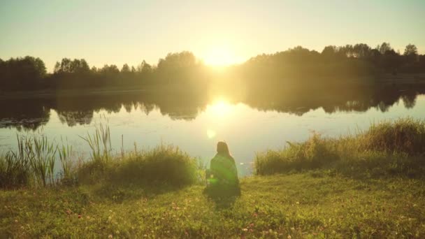 Lonely girl wrapped in plaid sitting on shore of lake and watching sunrise - Footage, Video