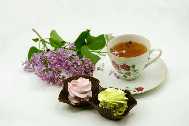 Se aíslan el té en la taza de porcelana, los malvaviscos rosados y amarillos, las flores de color lila púrpura sobre la superficie blanca
. - Foto, imagen