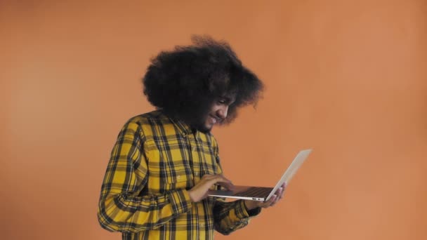 Afro-Amerikalı adam dizüstü bilgisayarda yazıyor ve turuncu arka planda gülümsüyor. Duyguların kavramı - Video, Çekim