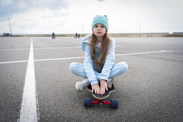 A girl in a blue hat and sweater sits on a large longboard along a large asphalt ground. - Zdjęcie, obraz