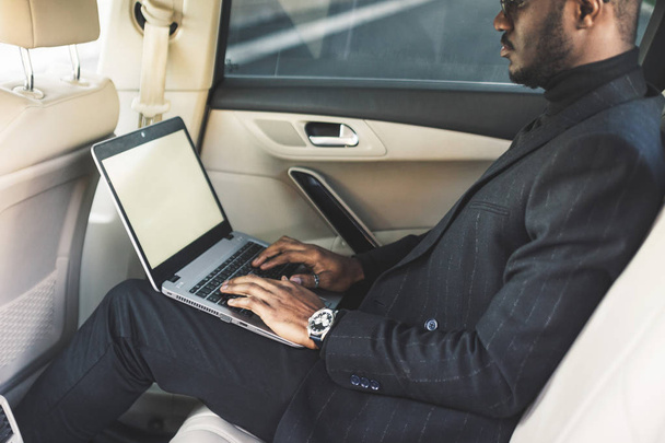 Άνθρωπος σε μια επαγγελματική στολή γράφουν σε φορητό υπολογιστή στο σαλόνι ενός ακριβό αυτοκίνητο με δέρμα εσωτερικό. - Φωτογραφία, εικόνα