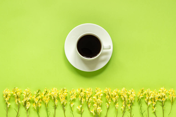 Cup of coffee and row yellow wild flowers Linaria on green paper background Flat lay Top view Concept Good morning or Hello spring Template for postcard, text or your design - Фото, изображение