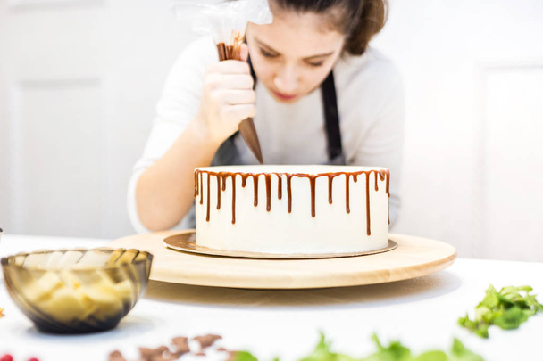 A confectioner squeezes liquid chocolate from a pastry bag onto a white cream biscuit cake on a wooden stand. The concept of homemade pastry, cooking cakes. - Photo, Image