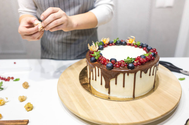 Confectioner decorates with berries a biscuit cake with white cream and chocolate. Cake stands on a wooden stand on a white table. The concept of homemade pastry, cooking cakes. - 写真・画像