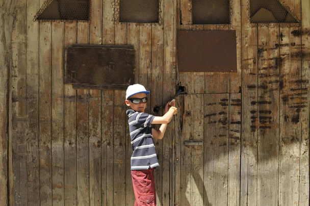 Garçon avec un bouclier et des lunettes de soleil essaie d'ouvrir une vieille porte de grange en bois
 - Photo, image