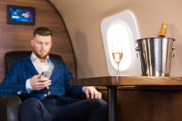A young successful businessman in an expensive suit sits in the chair of a private jet with a glass of champagne in his hand and looks out the window - Photo, image