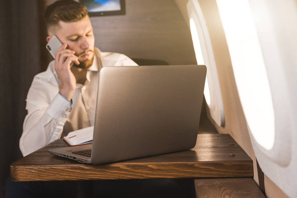 Young attractive and successful businessman talking on the phone and working while sitting in the chair of his private business plane - Foto, Imagem