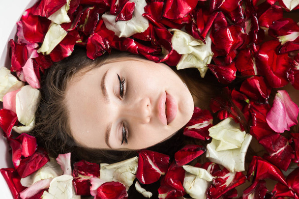 A young attractive girl takes a bathroom with flower petals and relaxes against the background of a beautiful light interior. Spa treatments for beauty and health with skin care - Fotó, kép