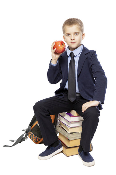 Schoolboy in a suit with an apple in his hand sitting on the books. Isolated on a white background.  - Фото, изображение