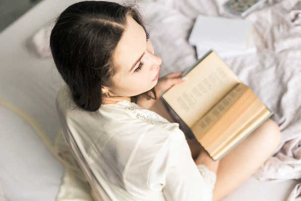 Attractive young girl in a nightgown sits in bed and reads a book - Fotó, kép