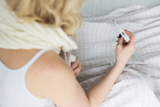 Beautiful blond woman sitting in bed with a thermometer and a scarf wrapped around the neck and a handkerchief. illness and health at home - Foto, afbeelding