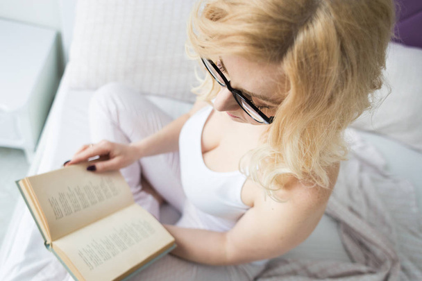 Attractive blonde woman in glasses with black rim sits on the edge of the bed at home with a book and reads - Photo, image