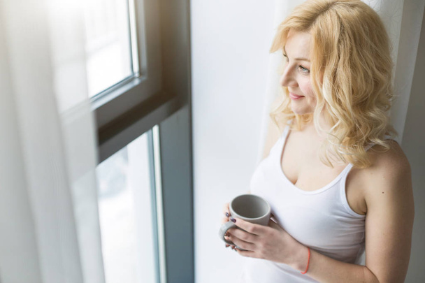 Egy vonzó szőke nő portréja áll az ablak mellett egy csésze kávéval vagy teával. - Fotó, kép
