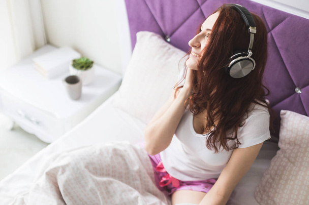 Attractive young brunette woman sits in the morning in her bed with headphones and a smartphone and listens to music in a good mood - Foto, imagen