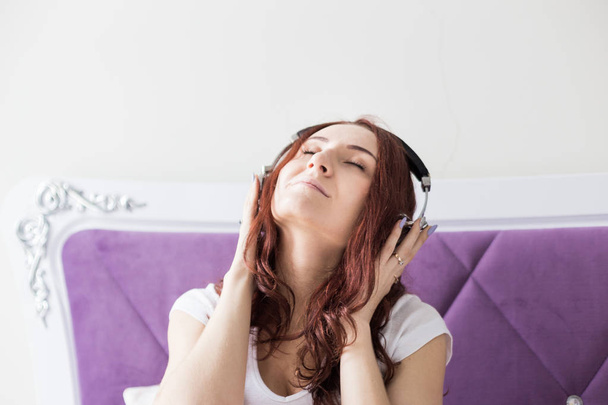 Attractive young brunette woman sits in the morning in her bed with headphones and a smartphone and listens to music in a good mood - Foto, Bild