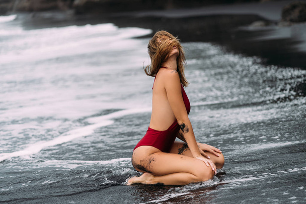 Sexy model with big breasts in a red swimsuit poses on black sand beach. - Foto, Imagem