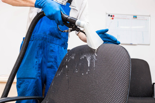 Young man in workwear and rubber gloves cleans the office chair with professional equipment. - Photo, image