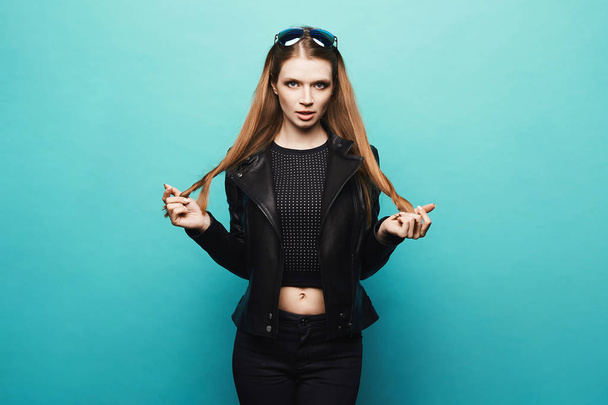 Sexy blonde model girl with a perfect body and with blue eyes in a stylish black leather jacket and in black jeans posing at the light blue background, isolated - Φωτογραφία, εικόνα