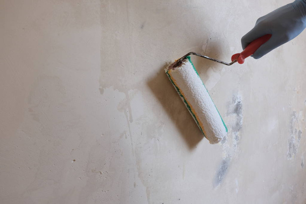 A male blue-gloved worker's hand applies a primer to a wall using a roller. Background of wet concrete. The concept of apartment renovation, finishing works, the preparation for painting. Copy space. - Photo, Image