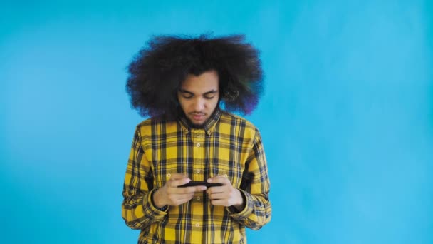 Hombre afroamericano jugando a juegos en Smartphone sobre fondo azul. Concepto de emociones
 - Metraje, vídeo