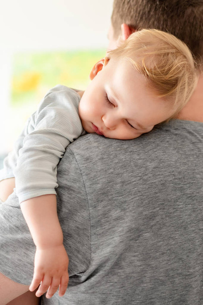 Portrait of cute adorable blond caucasian toddler boy sleeping on fathers shoulder indoors. Sweet little child feeling safety and care on daddys hand. Single father concept - Фото, изображение