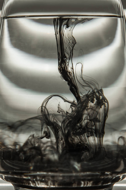 a cloud of ink in a transparent glass cup with clear water against the backdrop of a striped screen with rays of light. Template, layout, background, texture. macro diffusion black and white - Foto, Bild
