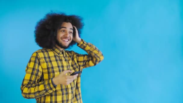 Feliz atraente jovem afro-americano usando telefone e recebendo boas notícias sobre fundo azul. Conceito de emoções
 - Filmagem, Vídeo