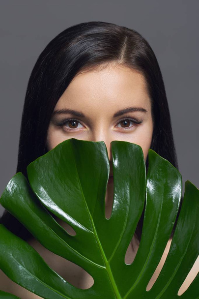 Studio beauty portrait of young brunette with natural make-up perfect skin with green exotic leaf on gray background. Concept of natural cosmetics, face and body care products. - Foto, Imagem