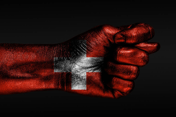 Una mano con una bandera de Suiza pintada muestra un higo, un signo de agresión, desacuerdo, una disputa sobre un fondo oscuro
. - Foto, imagen