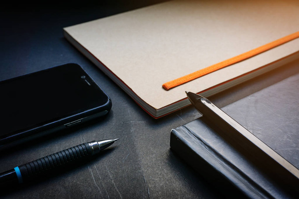 Zár megjelöl toll elhelyezett-ra fekete jegyzetfüzet, barna jegyzetfüzet, smartphone és ceruza-ra fekete iskolapad háttér-ban drámai gyújtás hangszín. Koncepció üzleti, tervezési vagy naplót. Nagy részére akármi használ. - Fotó, kép