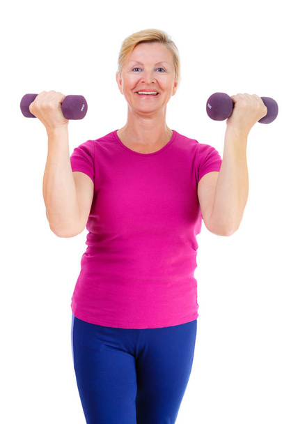 Pensioner woman sport. Old senior woman is engaged in inside sports. Woman looking at camera, holding violet dumbbells and cheerfully smiling while doing sport exercises. isolated on white background - Foto, Bild