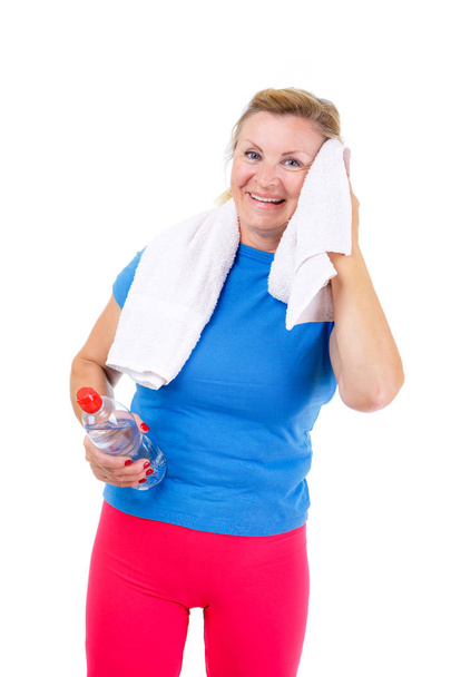 Seniorensport-Porträt. alte ältere blonde Frau steht isoliert auf weißem Hintergrund, lächelt und blickt in die Kamera mit Flasche Wasser, Handtuch um den Hals nach dem Sport-Training - Foto, Bild