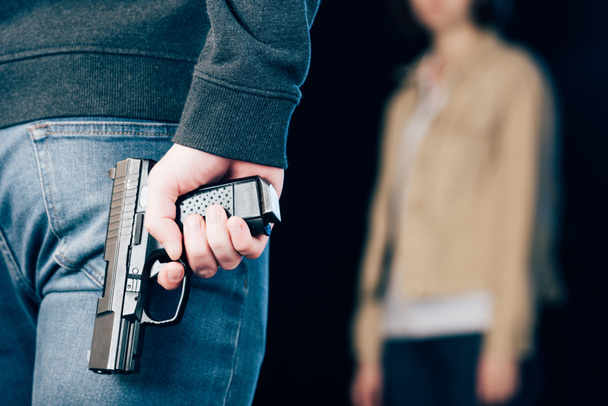 περικομμένη όψη του εγκληματικού κρυμμένο όπλο ενώ στέκεται κοντά γυναίκα σε μαύρο - Φωτογραφία, εικόνα