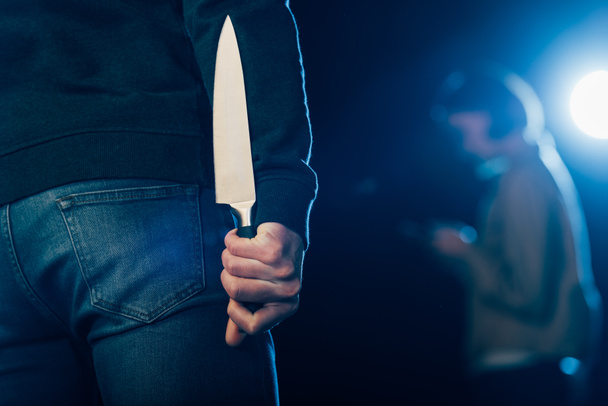 cropped view of murderer hiding knife behind back on black - Foto, Imagen