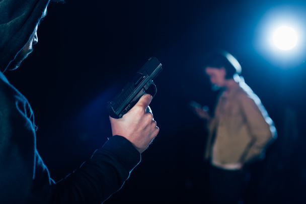 cropped view of killer in hood holding gun and looking at woman on black - Fotoğraf, Görsel