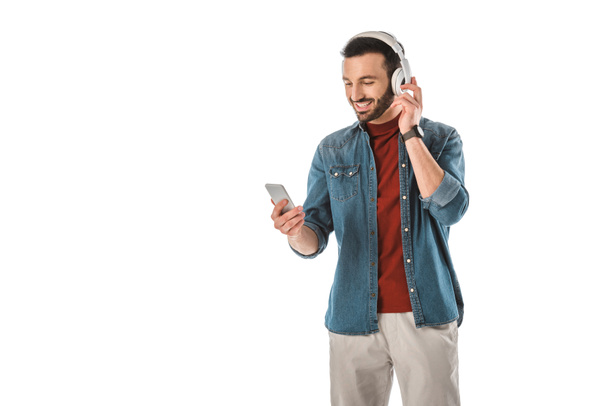 cheerful man listening music in headphones and using smartphone isolated on white - Fotó, kép